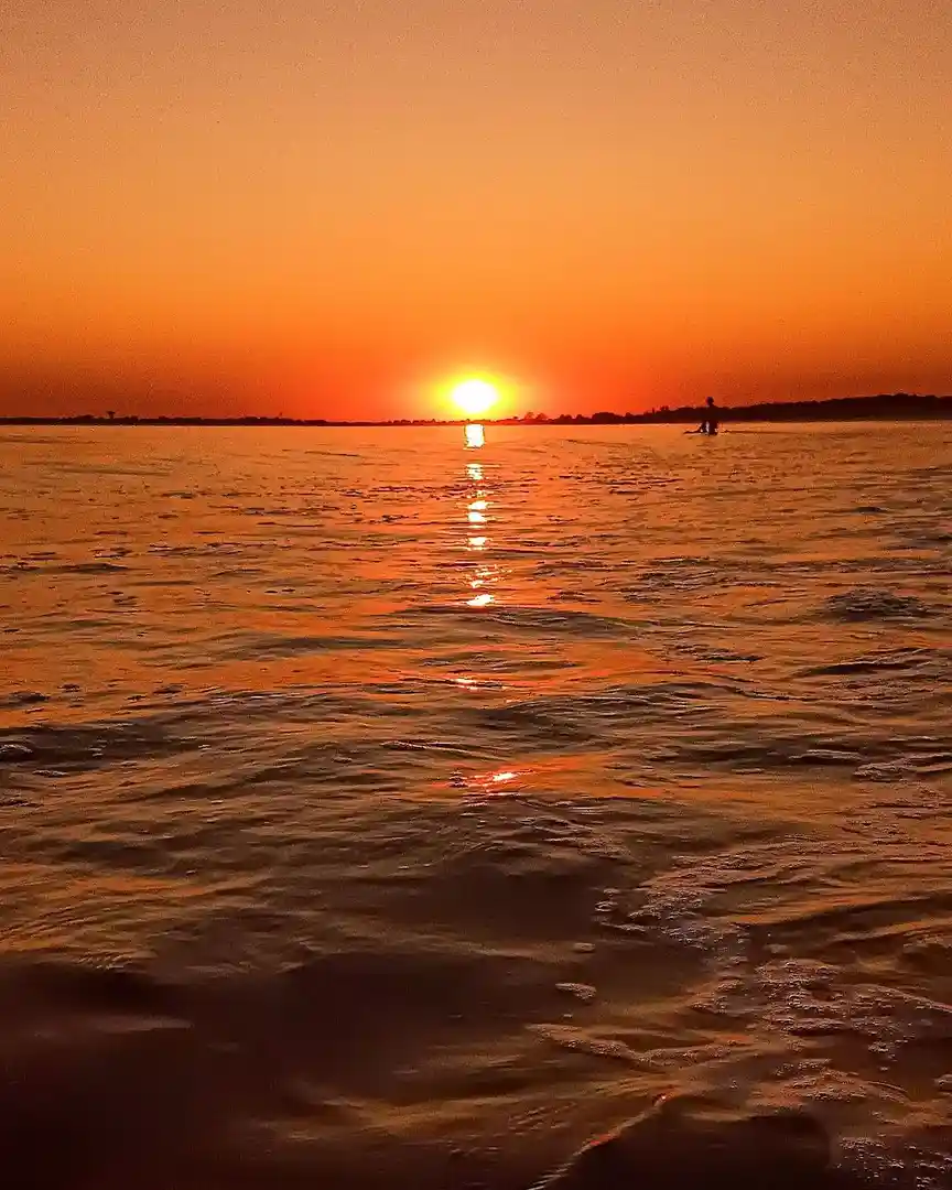 A serene sunset over water with a person silhouetted in the distance, the sun low on the horizon casting orange reflections…
