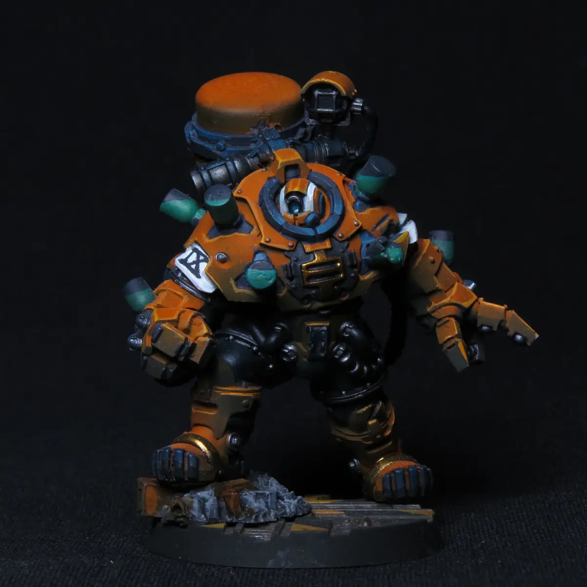 A heavily armored, orange and teal painted miniature figure stands on a rocky base against a black background. The figure…