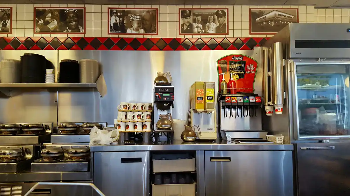 A photo of a Waffle House kitchen counter with stainless steel appliances, coffee machines, soda dispensers, and stacks of…