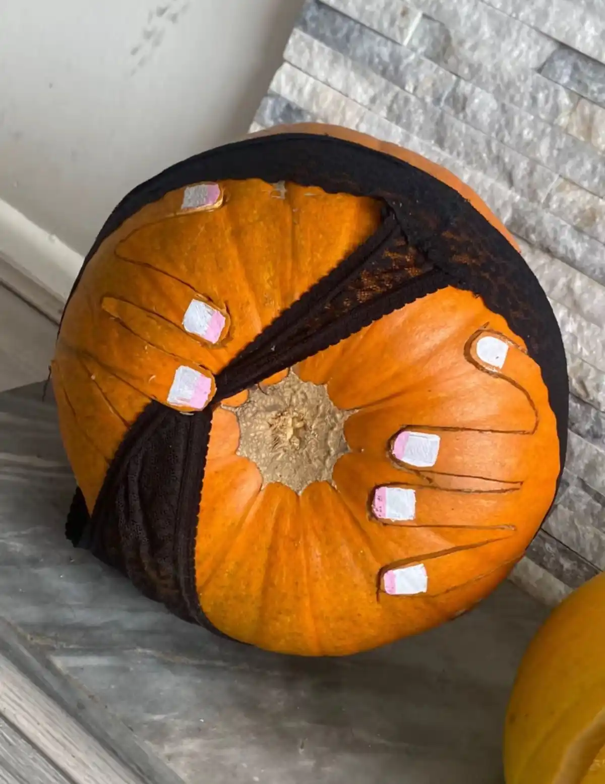 A close-up of an orange pumpkin with a black lace bra draped over its top half, featuring painted fingernails on its…