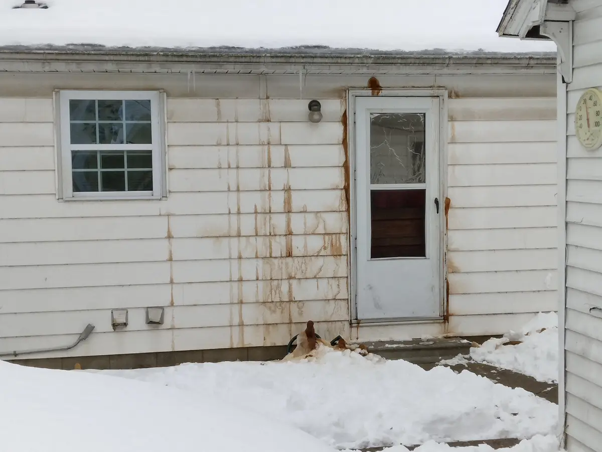A snowy exterior shot of a white-sided house with a cracked screen door and a window, showing brown water stains running…