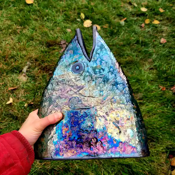 A hand holds up a large, triangular glass sculpture shaped like a fish head, featuring iridescent blue, purple, and gold…
