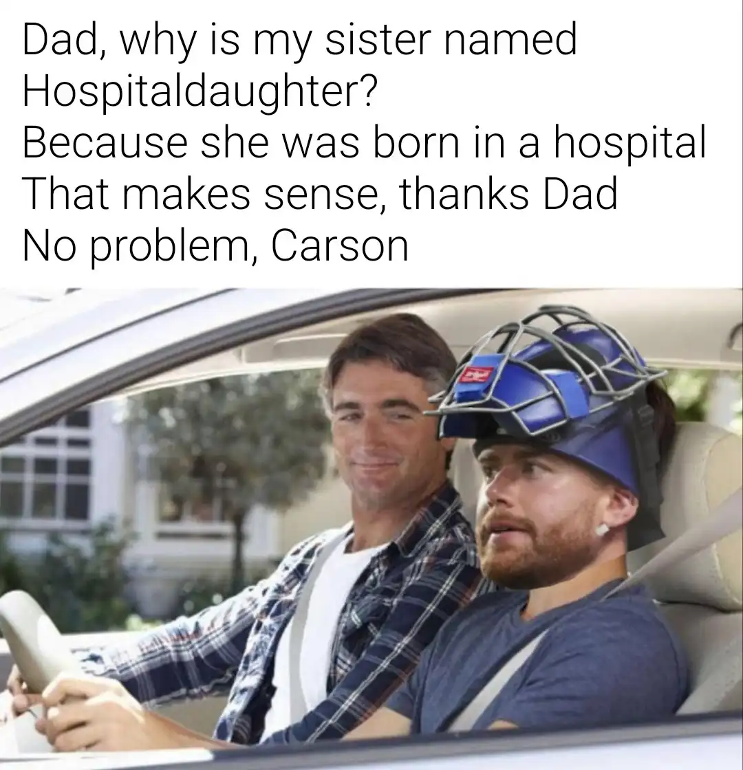A man in a plaid shirt drives a car while smiling, next to a passenger wearing a blue catcher's mask who looks confused. The…