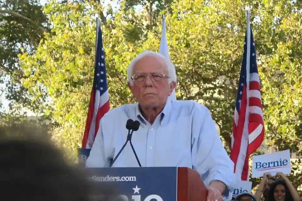 An older man with white hair and glasses stands at a podium with microphones, flanked by two American flags and a crowd…