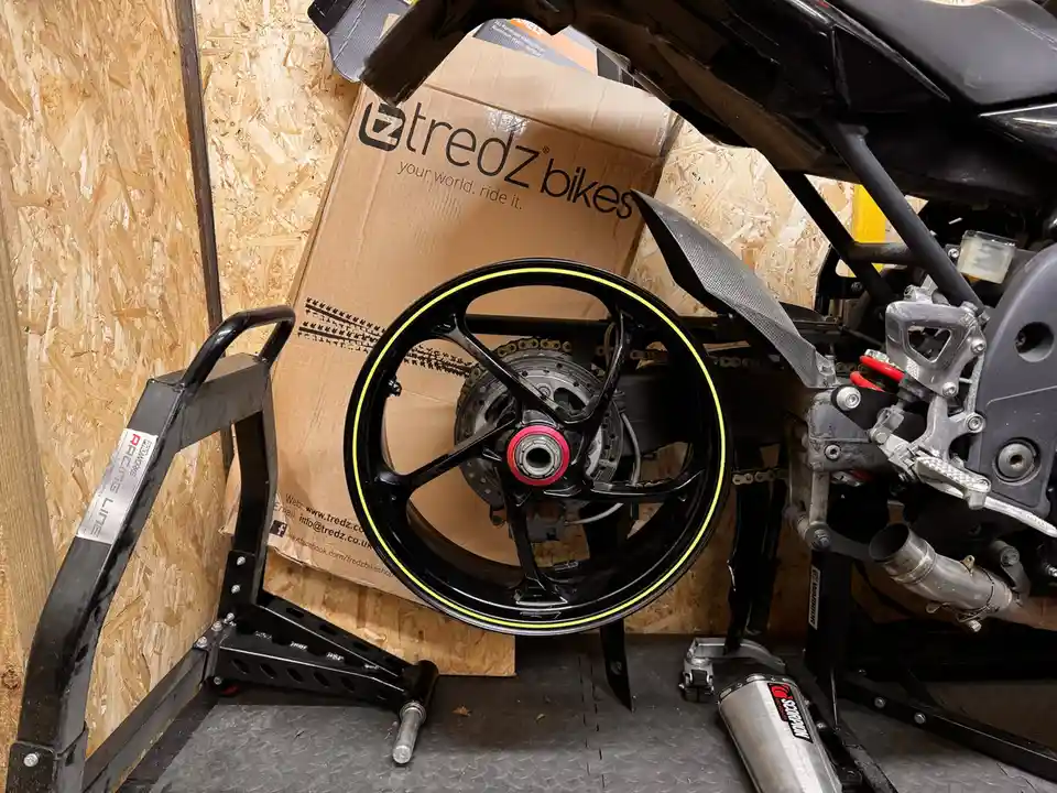 A close-up photo shows a black motorcycle wheel with a yellow rim stripe resting on a black stand in a garage. Behind it is…