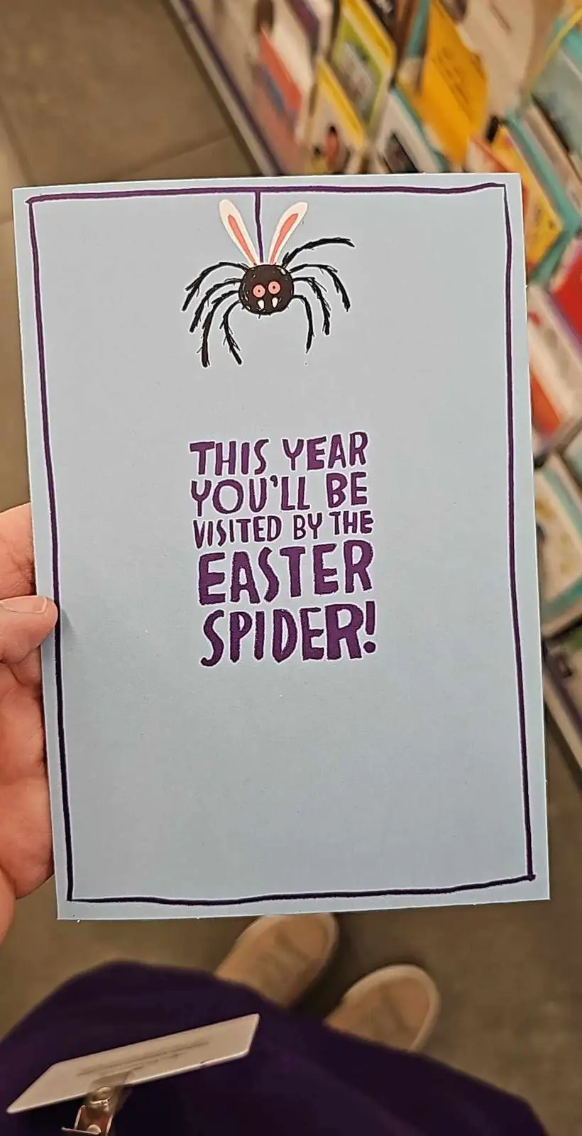 A hand holds a light blue greeting card with a purple border. At the top is a cartoon spider with bunny ears and a smiling…