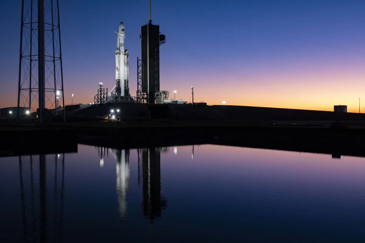 A SpaceX Starship rocket stands on a launchpad at dusk, illuminated by artificial lights against a gradient sky of blue and…