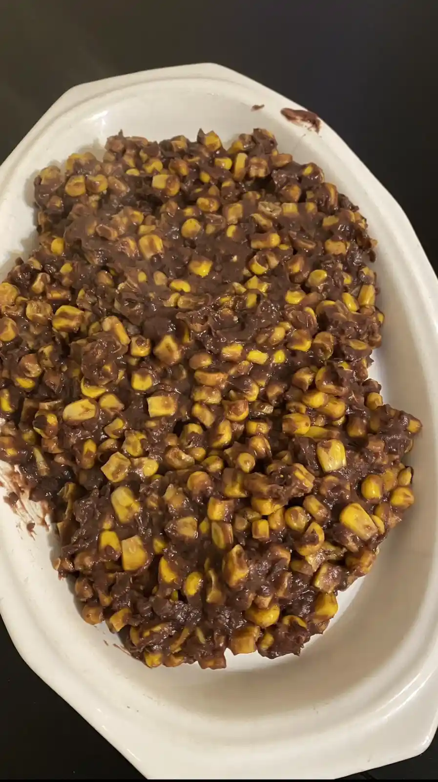 A white oval dish holds a mixture of yellow corn kernels coated in a thick, dark brown sauce that resembles melted…