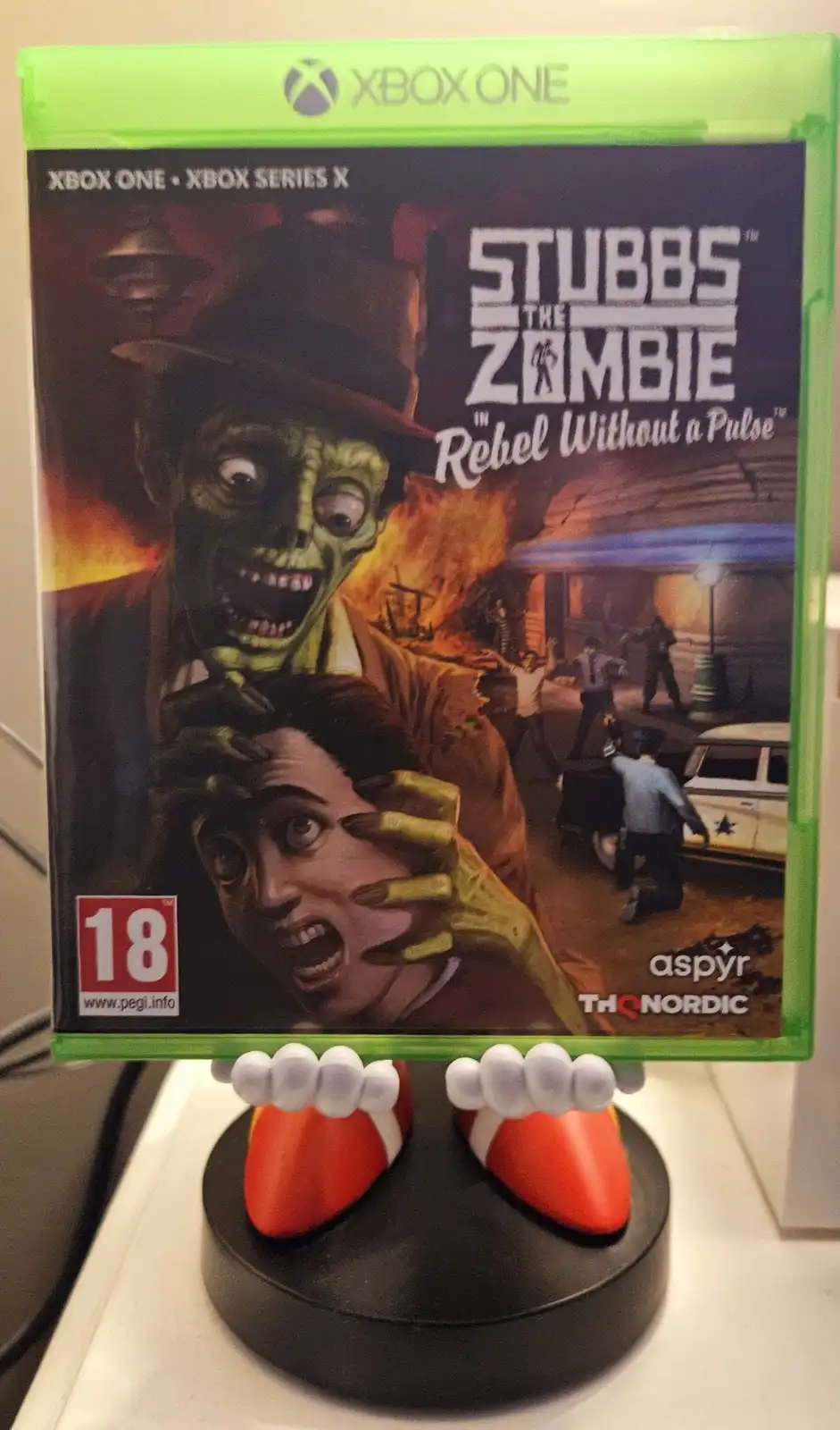A physical copy of the Xbox One game 'Stubbs the Zombie in Rebel Without a Pulse' is displayed upright on a stand shaped…
