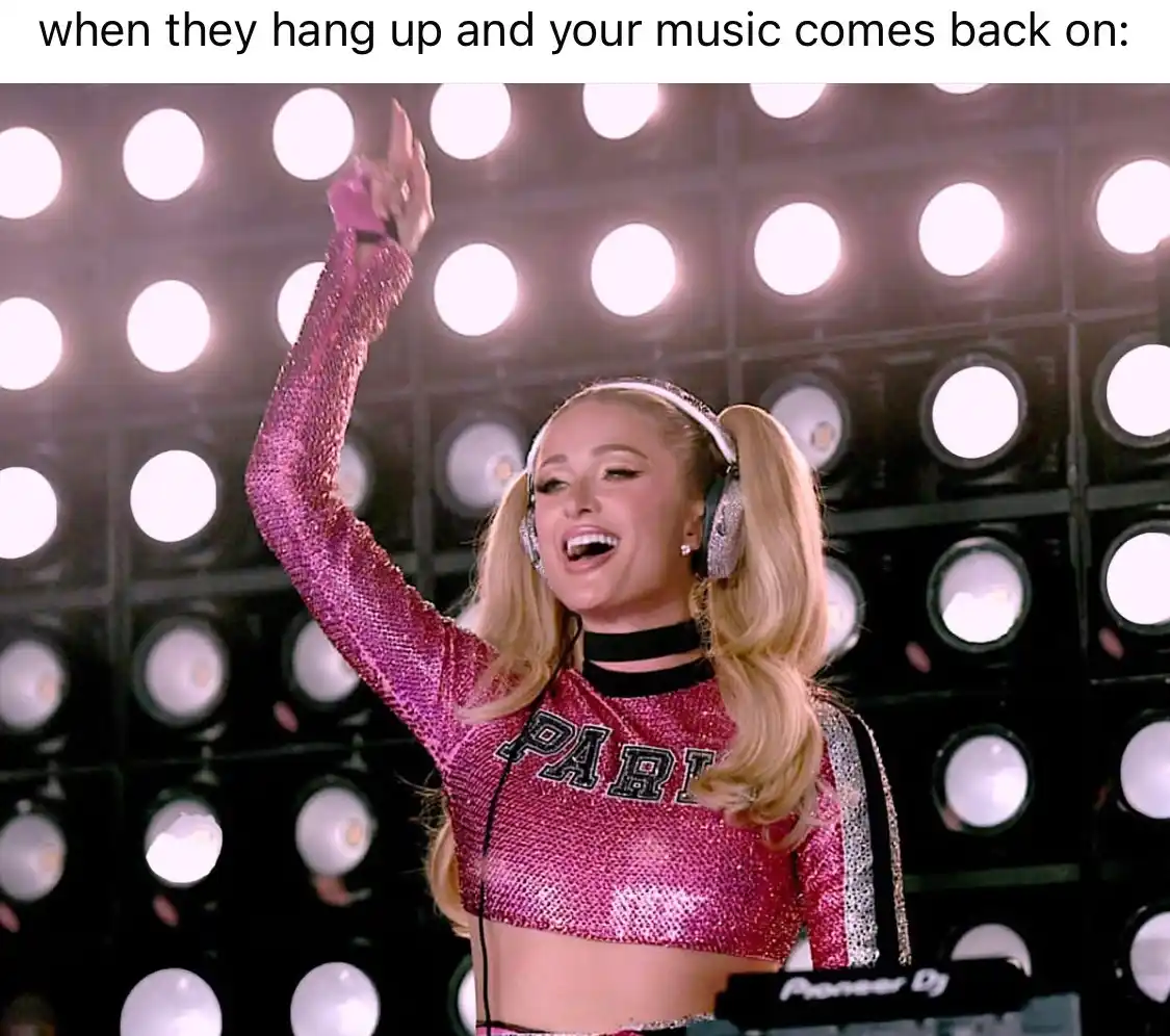 A woman with blonde pigtails and sparkly pink attire, wearing headphones and raising one hand in celebration, stands before…
