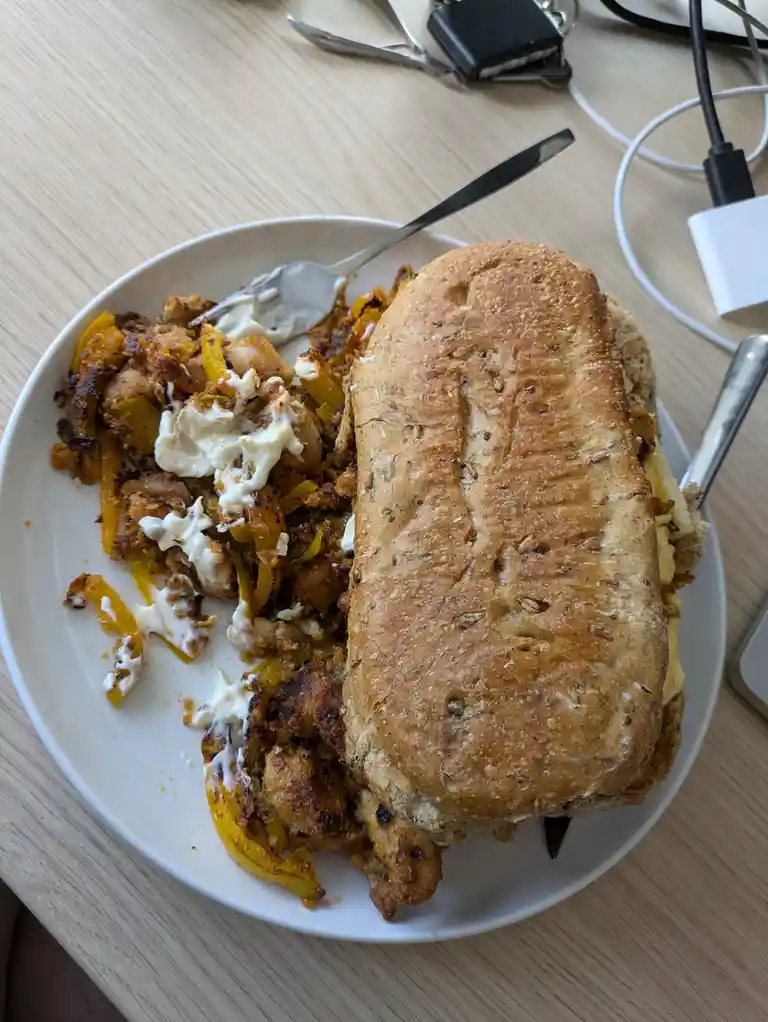 A partially eaten sandwich with a large, crusty roll sits on a white plate next to a pile of leftover chicken and yellow…