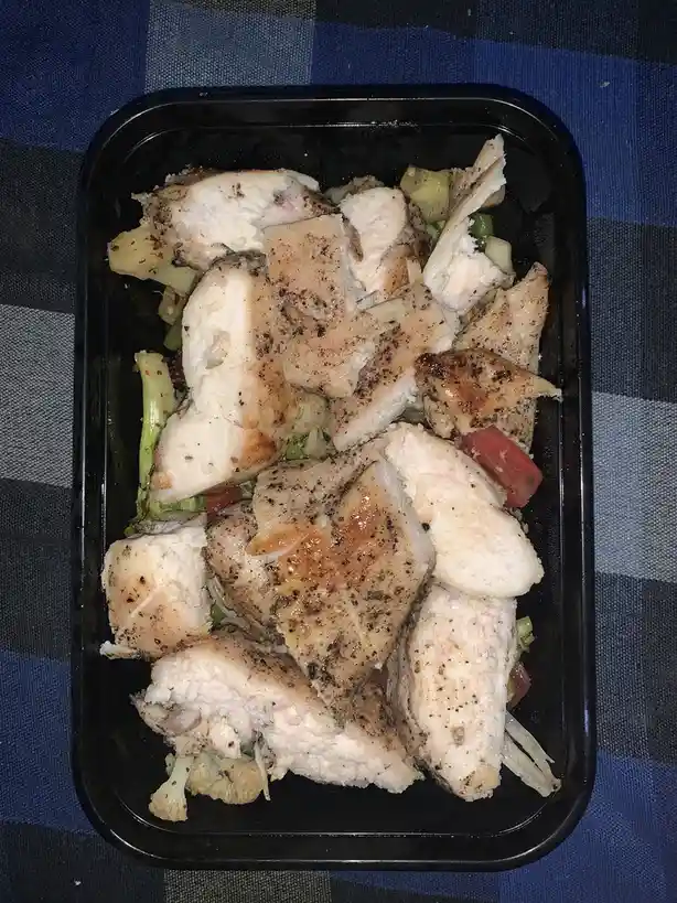 A black plastic container filled with chopped grilled chicken breast, mixed vegetables like broccoli and tomato, and…