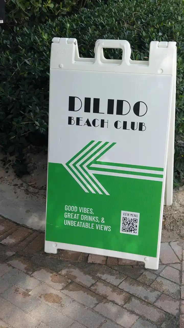 A white and green A-frame sign for 'DILIDO BEACH CLUB' stands on a brick sidewalk next to green bushes. The sign features a…