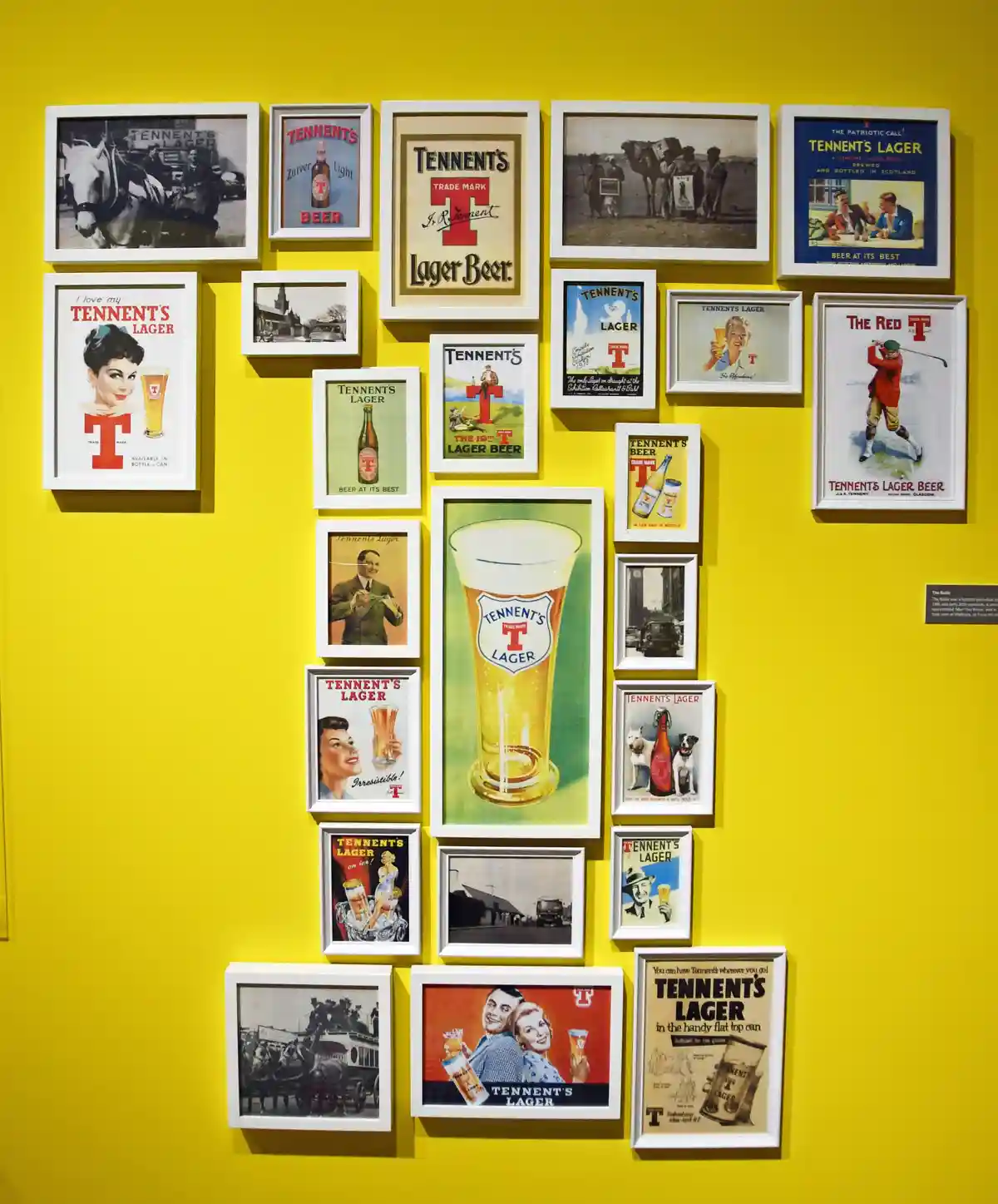 A bright yellow wall is covered with numerous framed vintage advertisements for Tennent's Lager beer, featuring various…