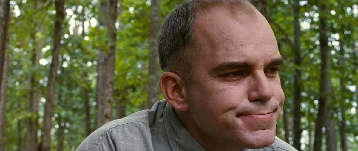 A man with short dark hair and a subtle, knowing smirk is shown in profile against a blurred forest background. He wears a…