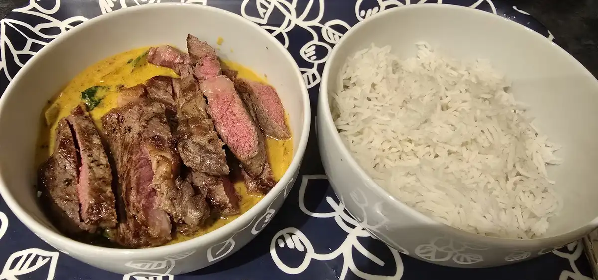 Two white bowls sit side-by-side on a dark blue patterned tablecloth. The left bowl contains sliced medium-rare steak in a…