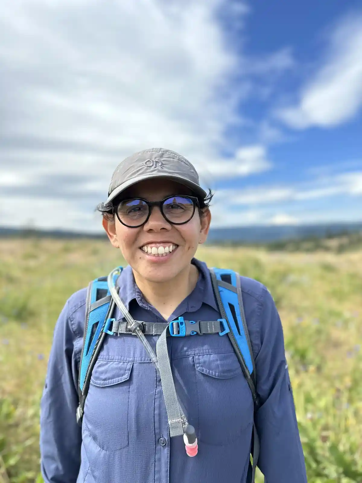 A smiling woman wearing glasses, a grey cap, and a blue hiking shirt with a hydration pack stands in a grassy field under a…