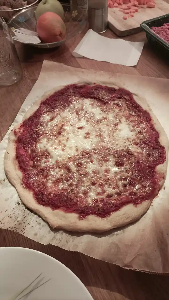 A homemade pizza sits on parchment paper on a wooden table, featuring a thick red sauce layer around the crust and a central…