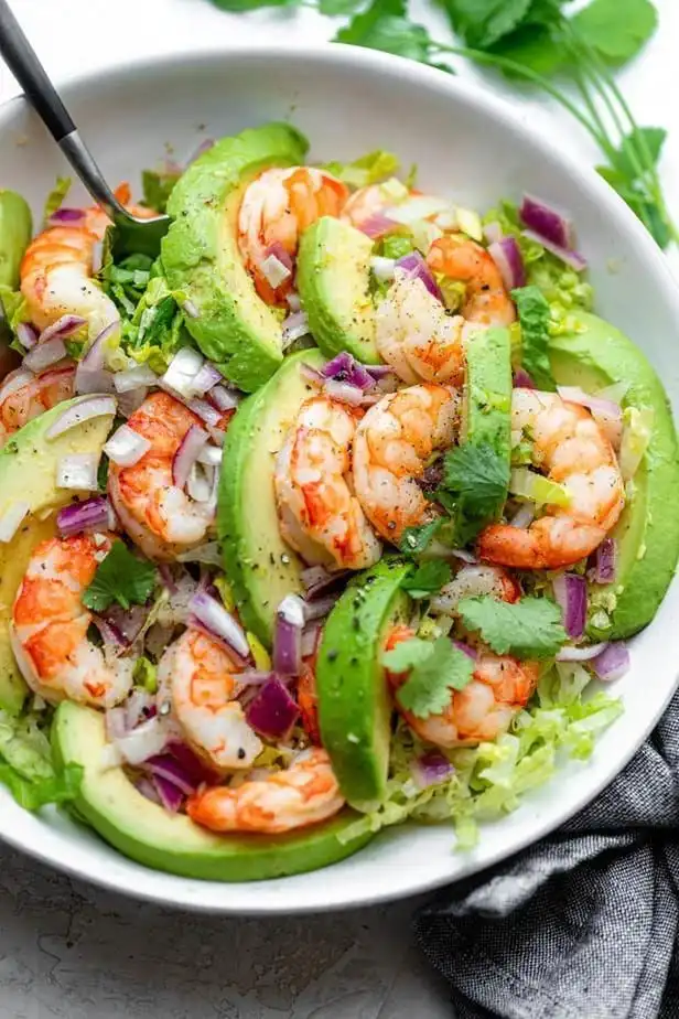 A white bowl filled with a vibrant shrimp avocado salad, featuring cooked pink shrimp, thick avocado slices, chopped red…