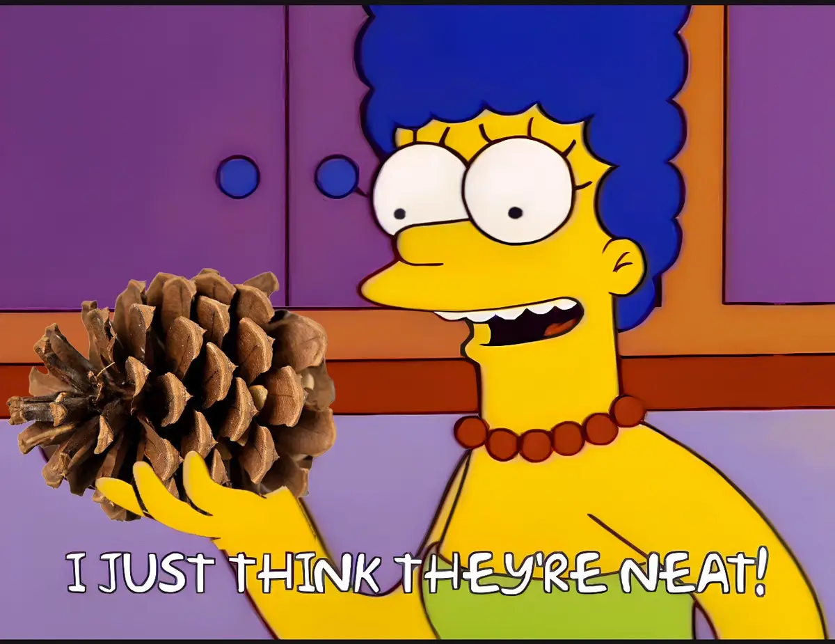 Marge Simpson holds a large pinecone with a wide-eyed, enthusiastic expression while standing in front of purple cabinets…