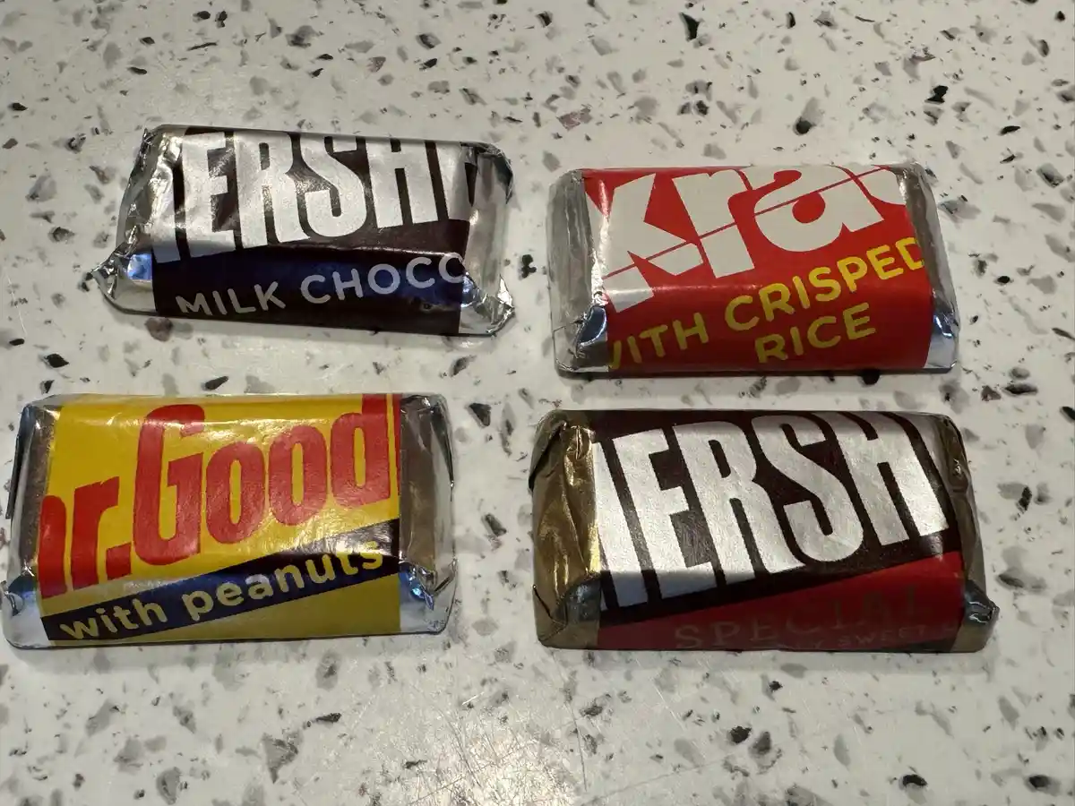 Four chocolate bars are arranged on a speckled countertop: a Hershey's Milk Chocolate, a KitKat with Crisped Rice, a Mr…