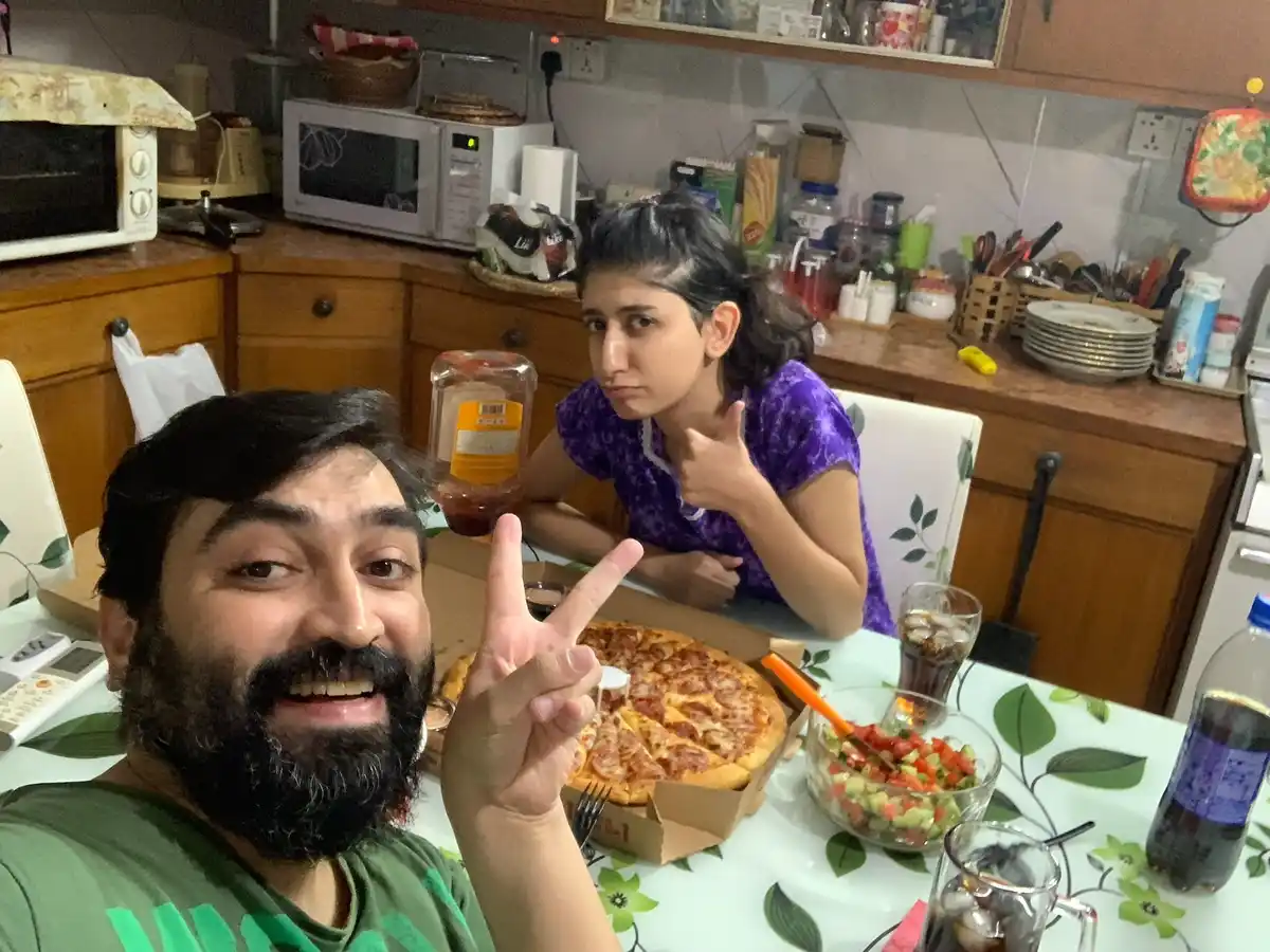 A man with a beard smiles and makes a peace sign at the camera while sitting at a kitchen table with a pizza in front of…