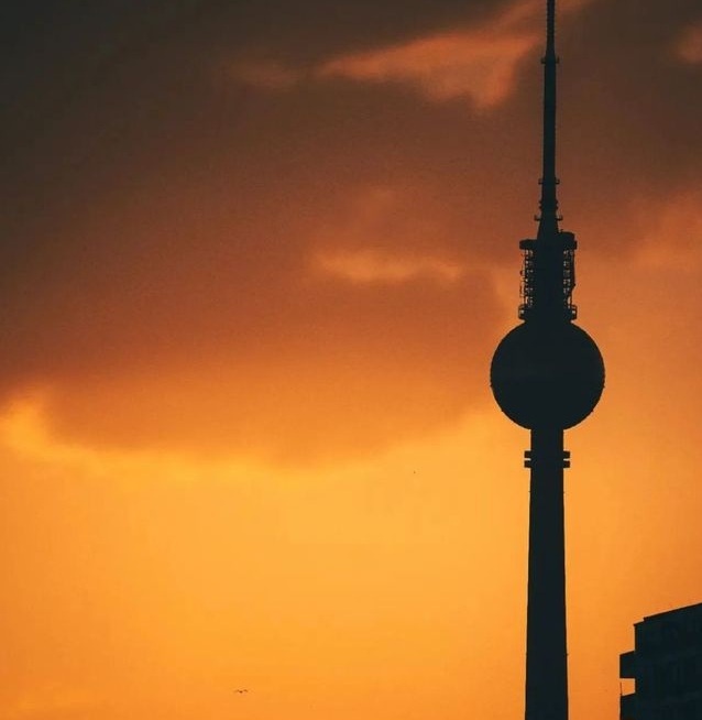 A silhouette of the Berlin TV Tower against a vibrant orange and dark orange sunset sky. The tower's distinctive spherical…
