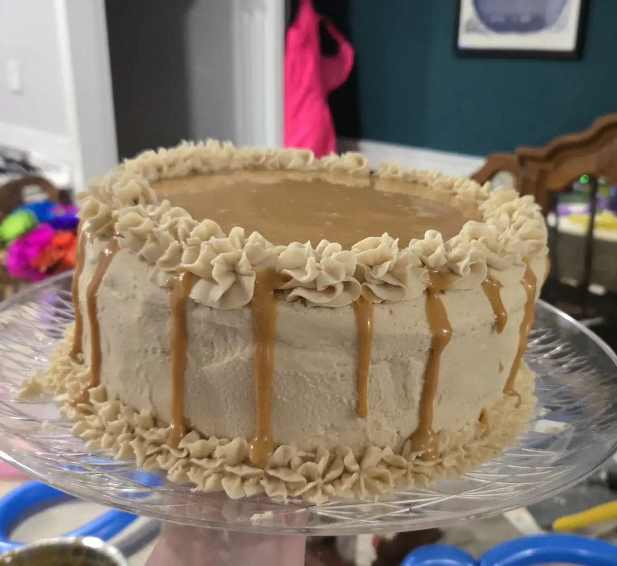 A round cake with light brown frosting, piped rosettes around the top and bottom edges, and caramel sauce dripping down the…