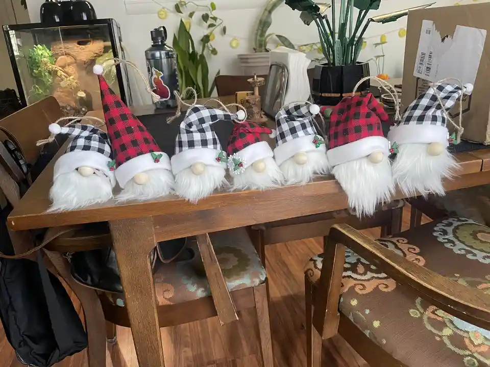 Six identical decorative gnomes with white beards and wooden noses sit in a row on a wooden dining table. They wear…