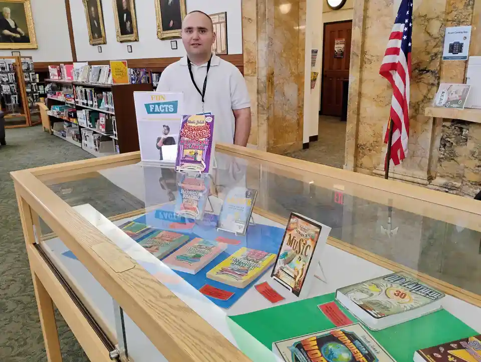 A man in a white polo shirt stands behind a glass display case in a library, looking surprised or confused. The case…
