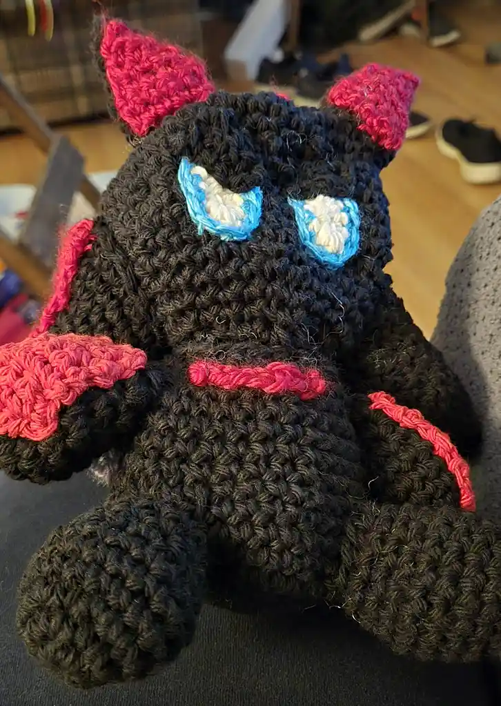 A handmade black crocheted stuffed animal resembling a cat with red crocheted horns, wings, and a collar, and blue-and-white…