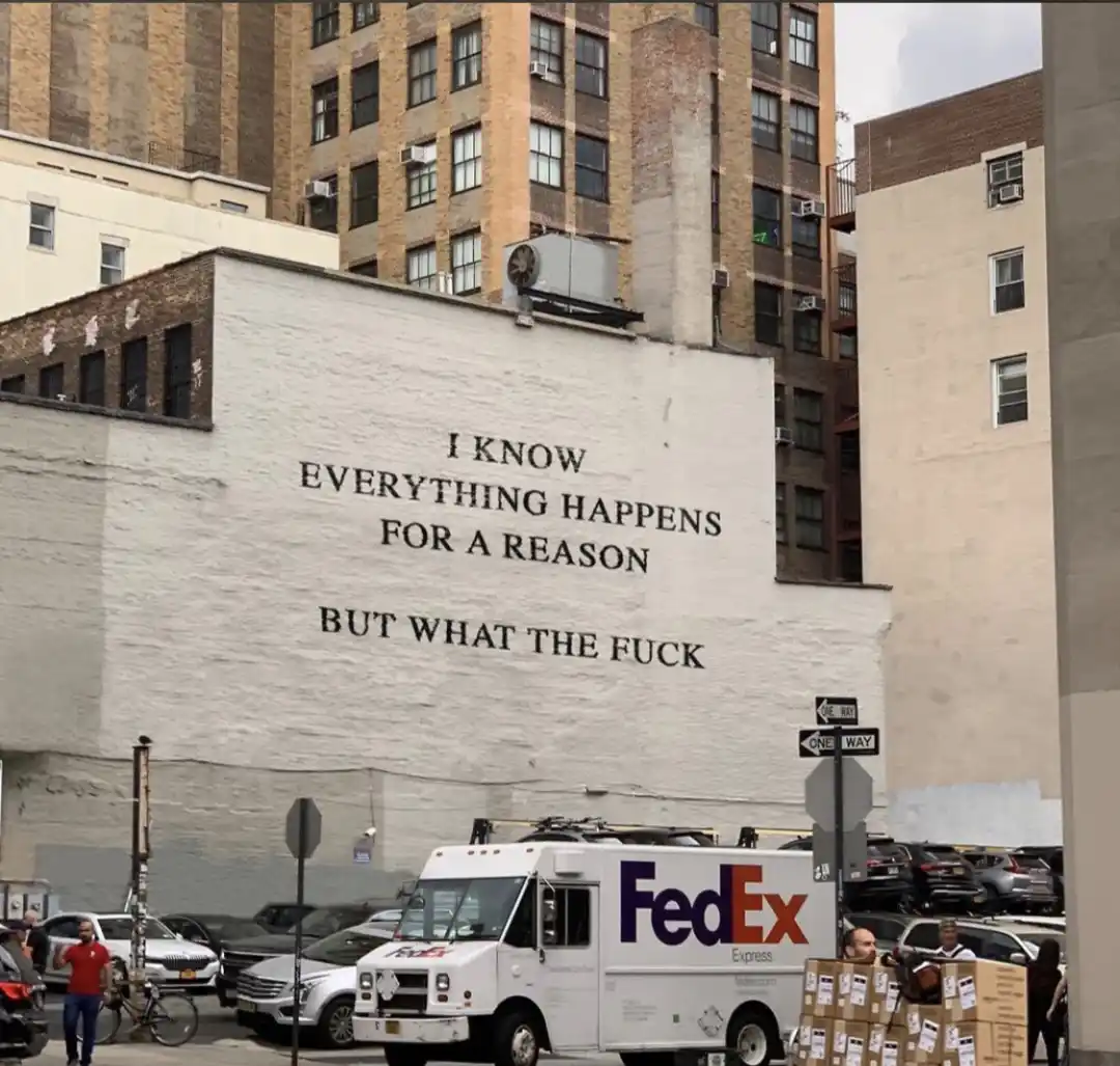 A large white wall in an urban setting has the text 'I KNOW EVERYTHING HAPPENS FOR A REASON BUT WHAT THE FUCK' painted on it…