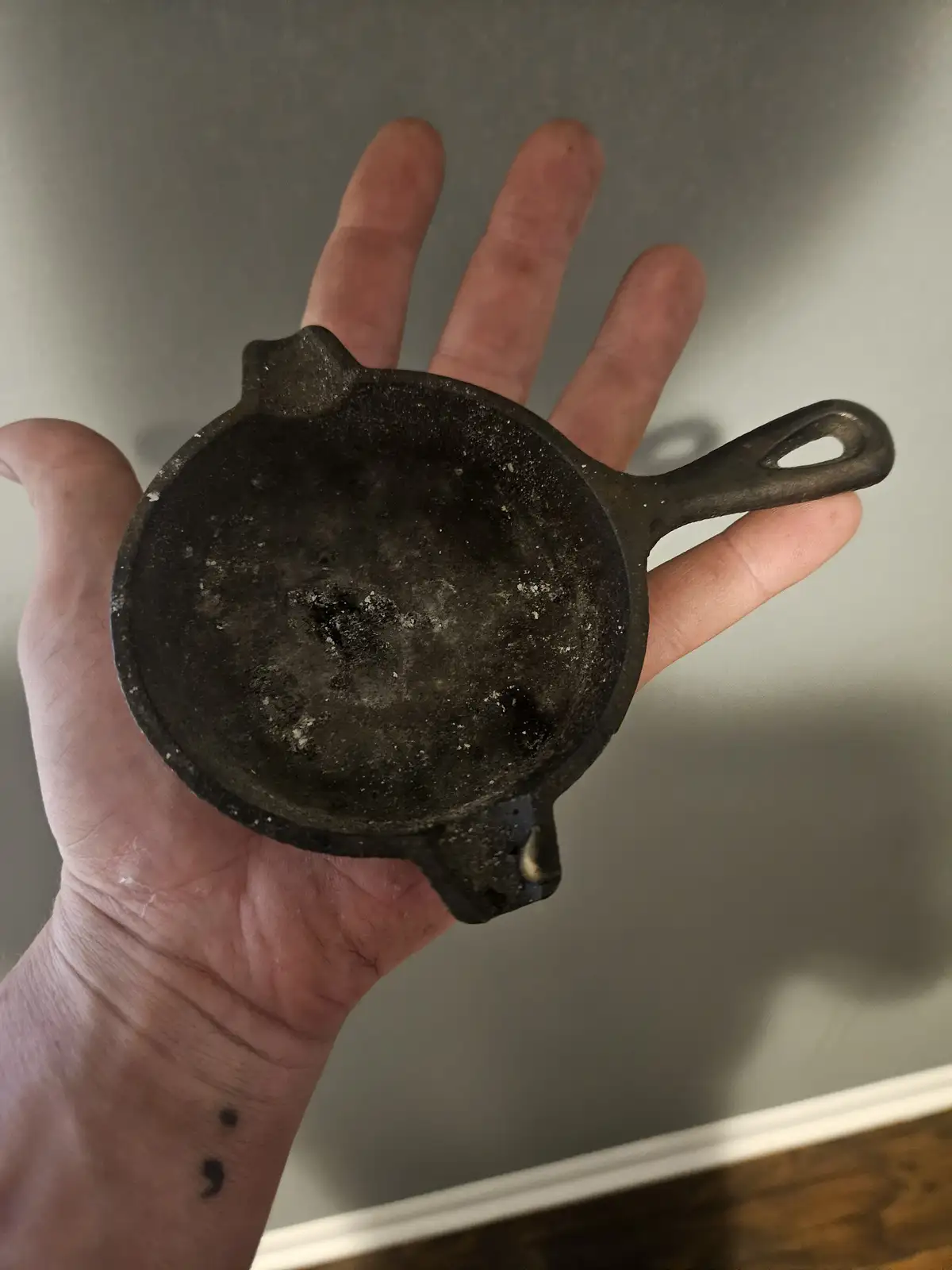 A person holds a very small, dark, well-used cast iron skillet in their open palm, showing its tiny size relative to their…