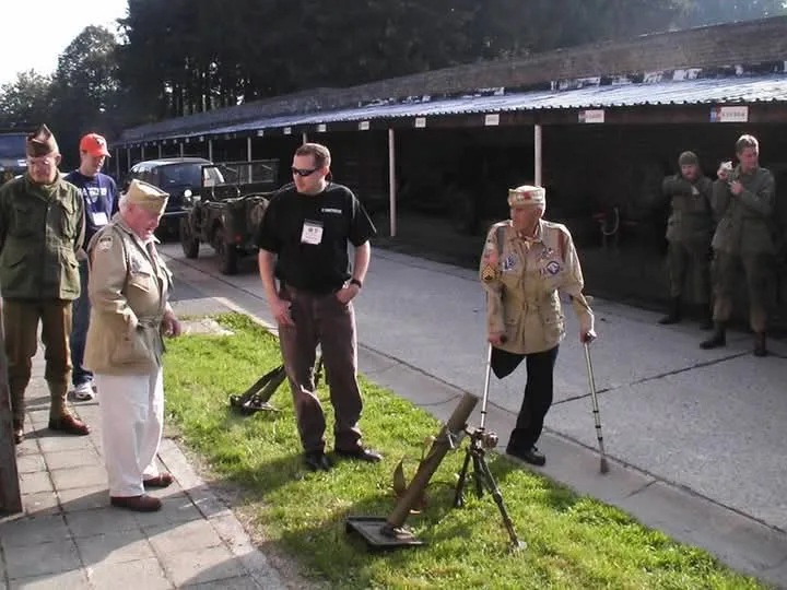 A group of men dressed in military-style uniforms stand outdoors near a shooting range. One older man in a khaki uniform and…