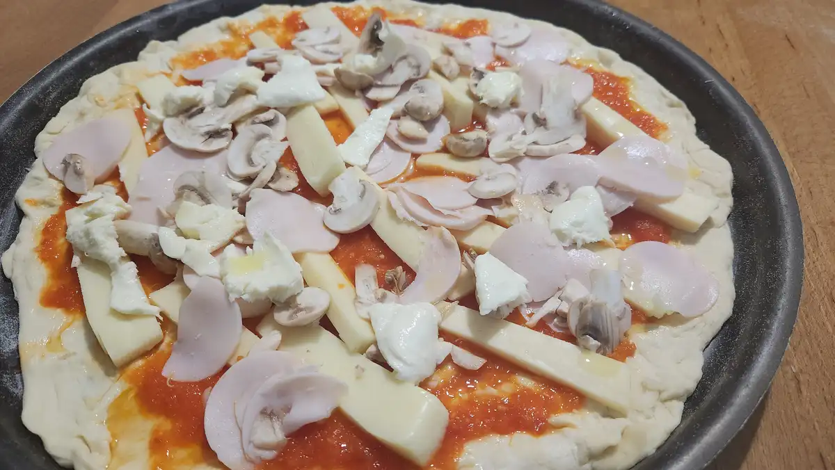 A close-up photo of an uncooked pizza in a black pan, covered with tomato sauce, sliced mushrooms, ham, and multiple types…