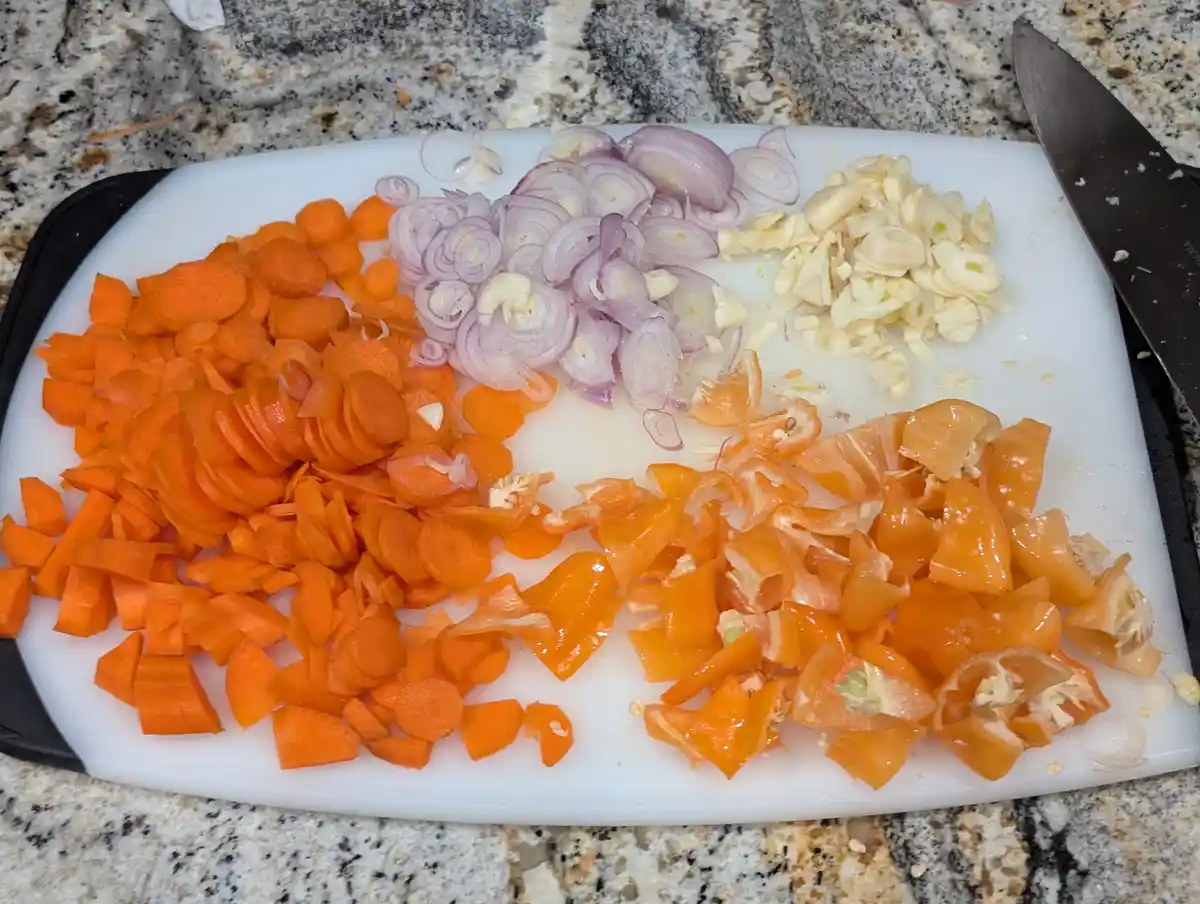 A white cutting board on a granite countertop holds neatly chopped vegetables: sliced carrots, chopped red onions, minced…