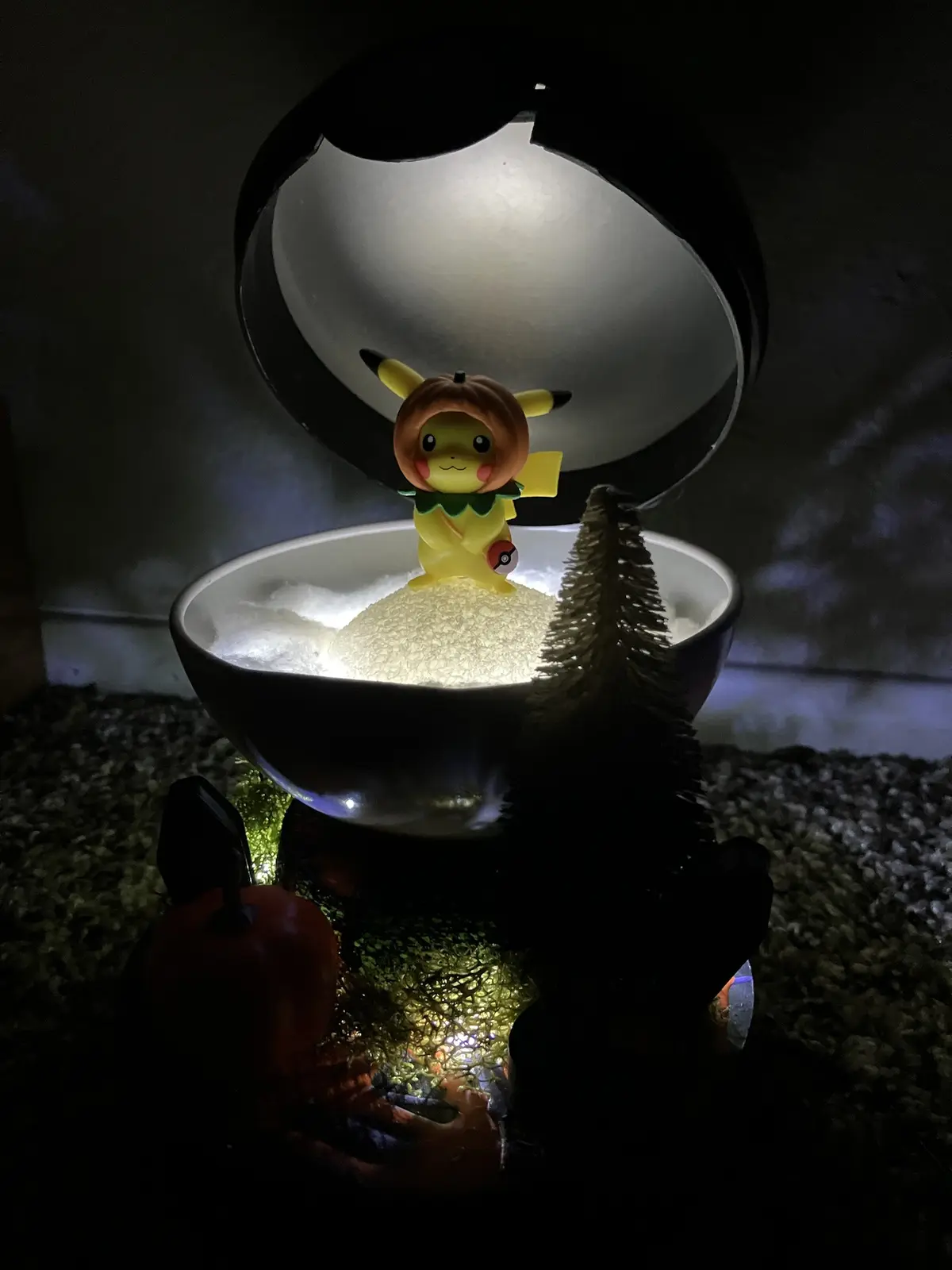 A yellow Pikachu figurine wearing a pumpkin hat sits inside a glowing, cracked Poké Ball-shaped bowl filled with white foam…
