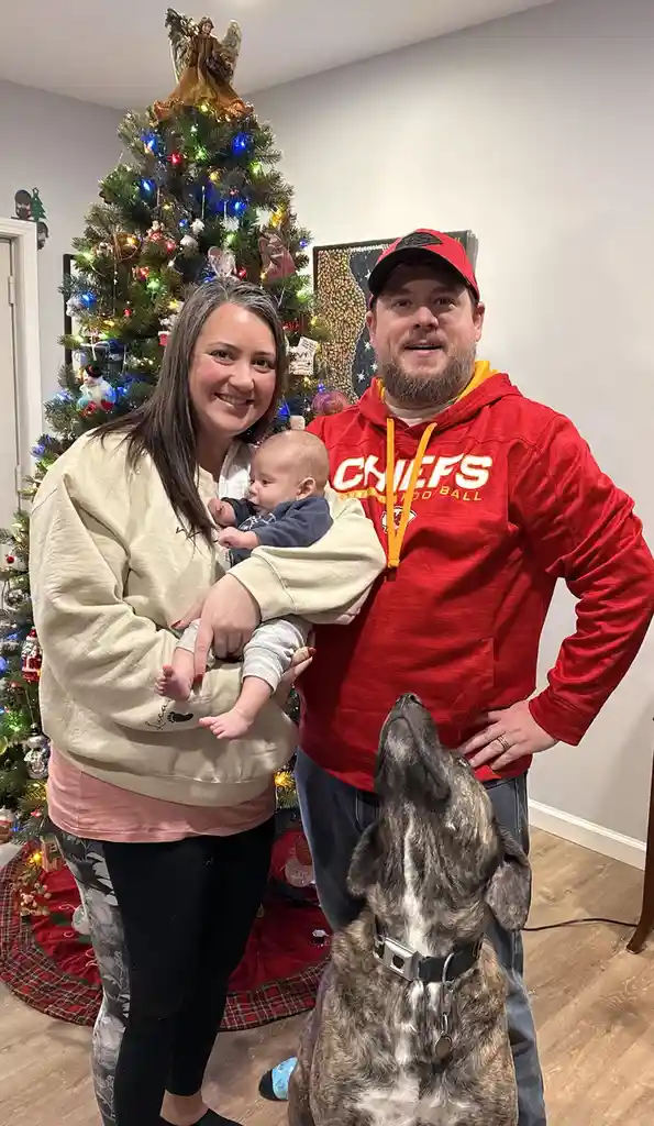 A family photo shows a woman holding a baby, standing next to a man in a red Chiefs hoodie, with a dog looking up at them. A…