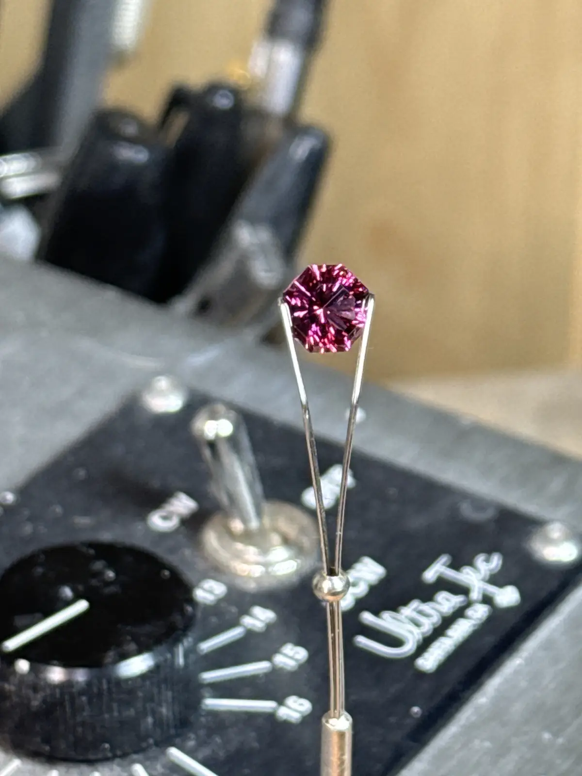A close-up photo shows a bright pink, octagon-cut gemstone held by a jeweler's tweezer over a black jeweler's loupe with…