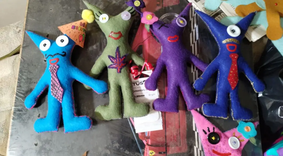 Four handmade felt dolls with cone-shaped heads and button eyes are arranged on a cluttered surface. The blue doll on the…