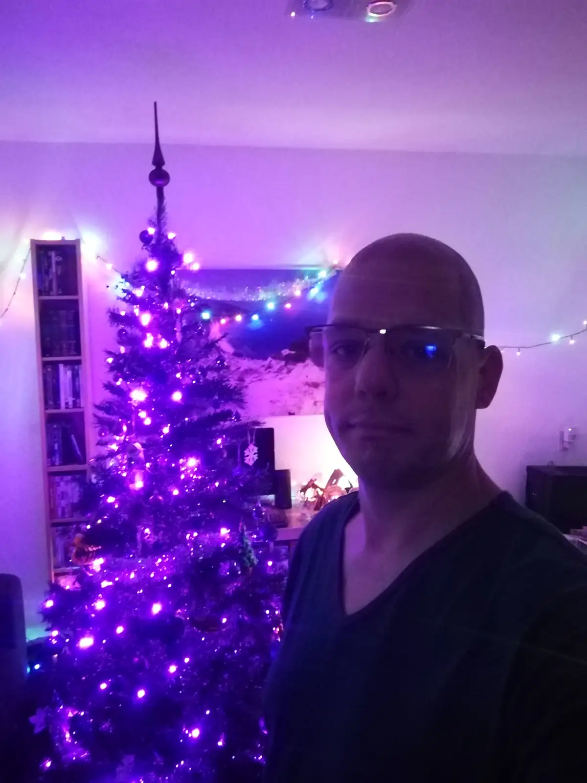 A bald man with glasses stands in front of a Christmas tree illuminated with purple lights, looking directly at the camera…