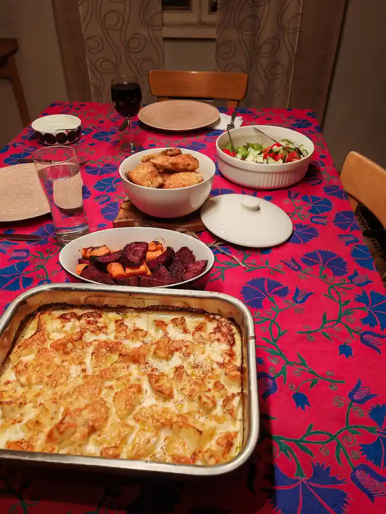 A dining table set with a vibrant red and blue floral tablecloth, featuring a large metal baking dish with a golden-brown…