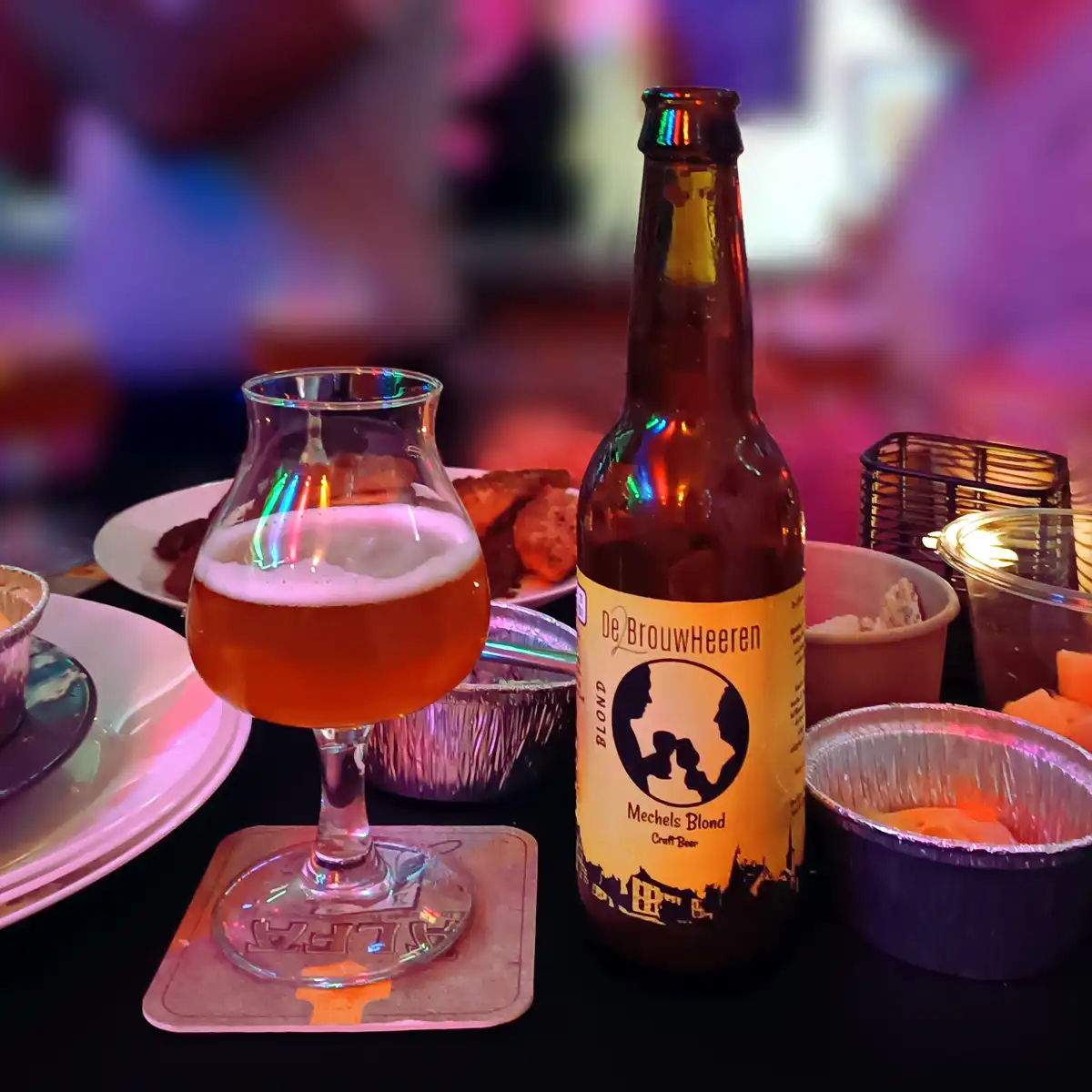 A close-up photo of a bottle of De BrouwHeeren Mechels Blond craft beer next to a tulip-shaped glass filled with the beer…