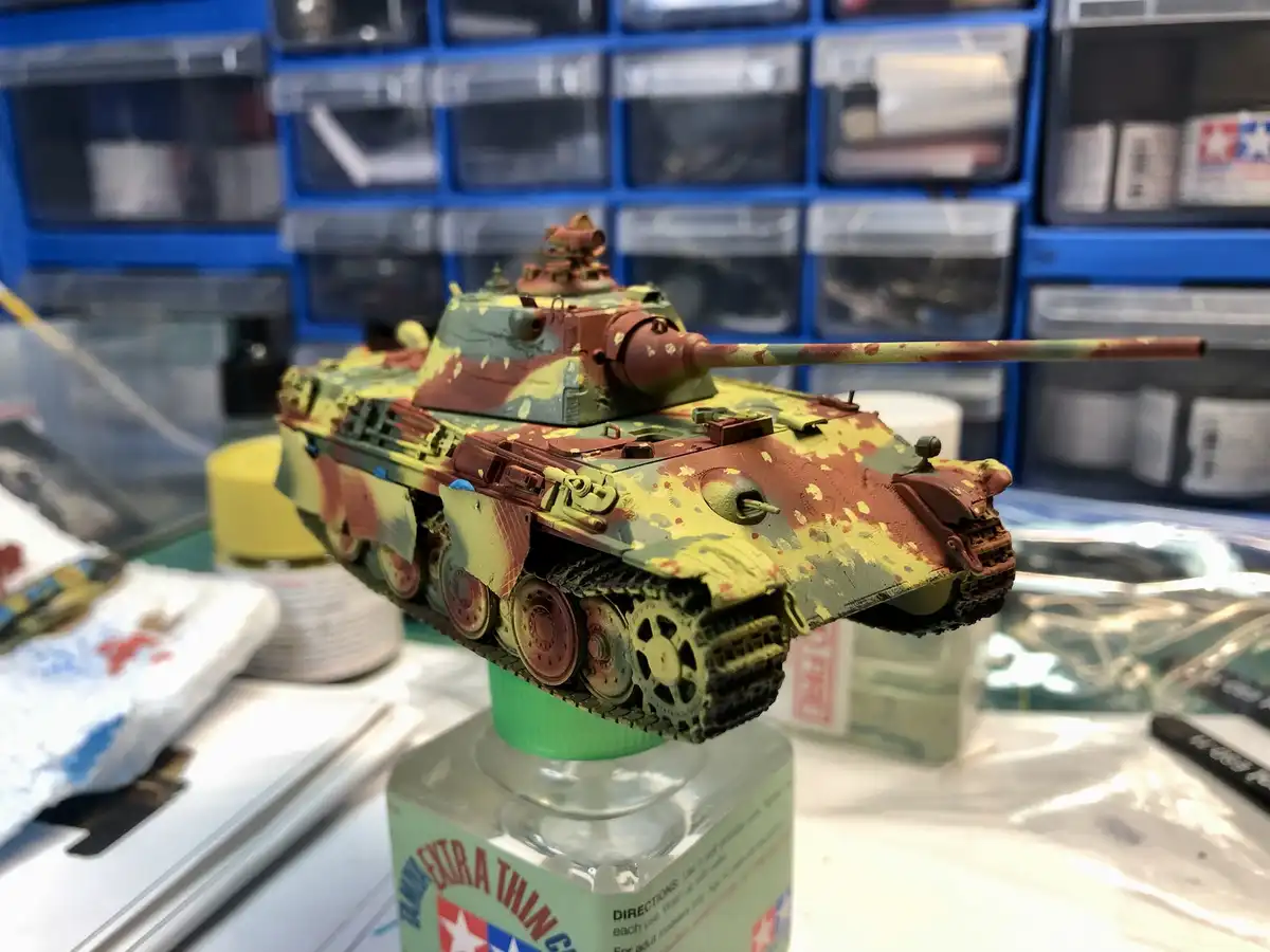 A scale model of a tank with messy, splattered camouflage paint sits on top of a bottle of Tamiya Extra Thin Clear…