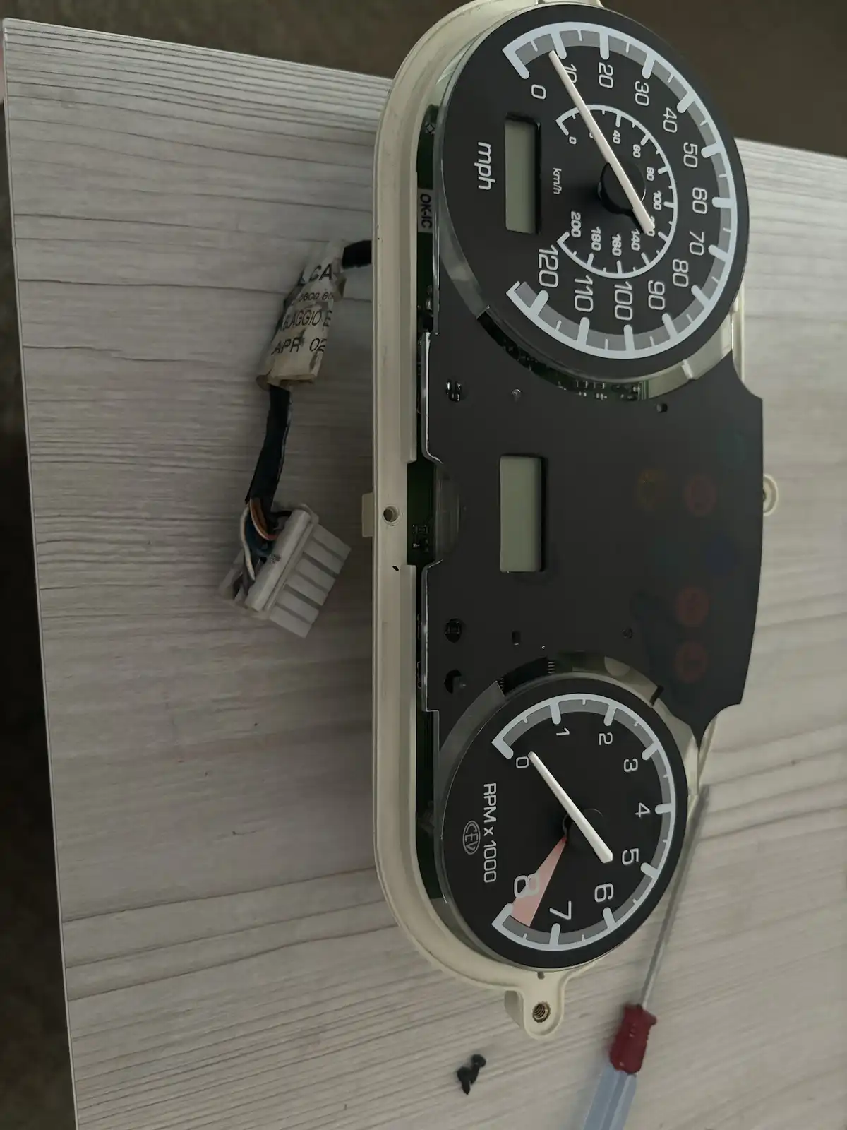 A disassembled car dashboard with a speedometer and tachometer, both needles pointing to zero, sits on a wooden surface next…