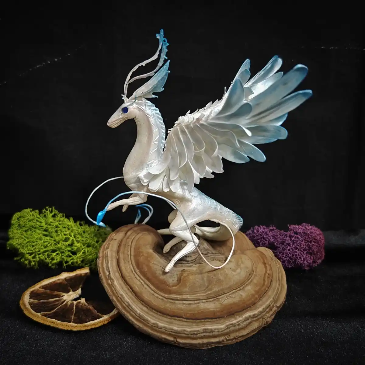 A white, winged, deer-like mythical creature with blue eyes and translucent blue wings stands on a large, layered brown…