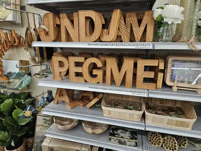 A store shelf displays large wooden letters arranged to spell 'JNDAM' and 'PEGME' on upper shelves, with a sign below each…