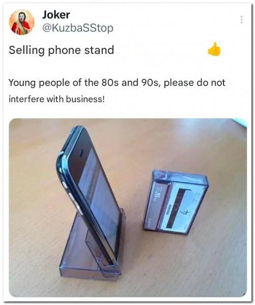 A smartphone is propped up on a clear plastic stand shaped like a cassette tape case, sitting on a wooden surface next to an…