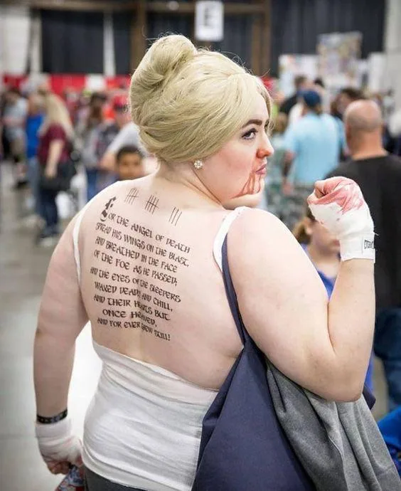 A woman at a convention with blonde hair in a bun, wearing a white tank top and carrying a bag, has a full back tattoo of a…