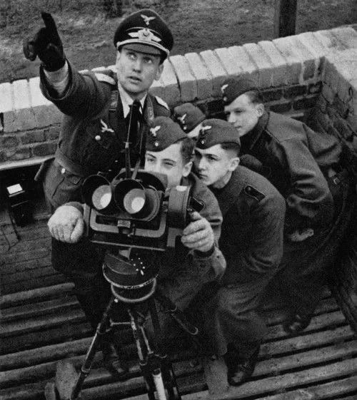 A black-and-white historical photo shows four men in Nazi military uniforms on a wooden platform, one pointing forward while…